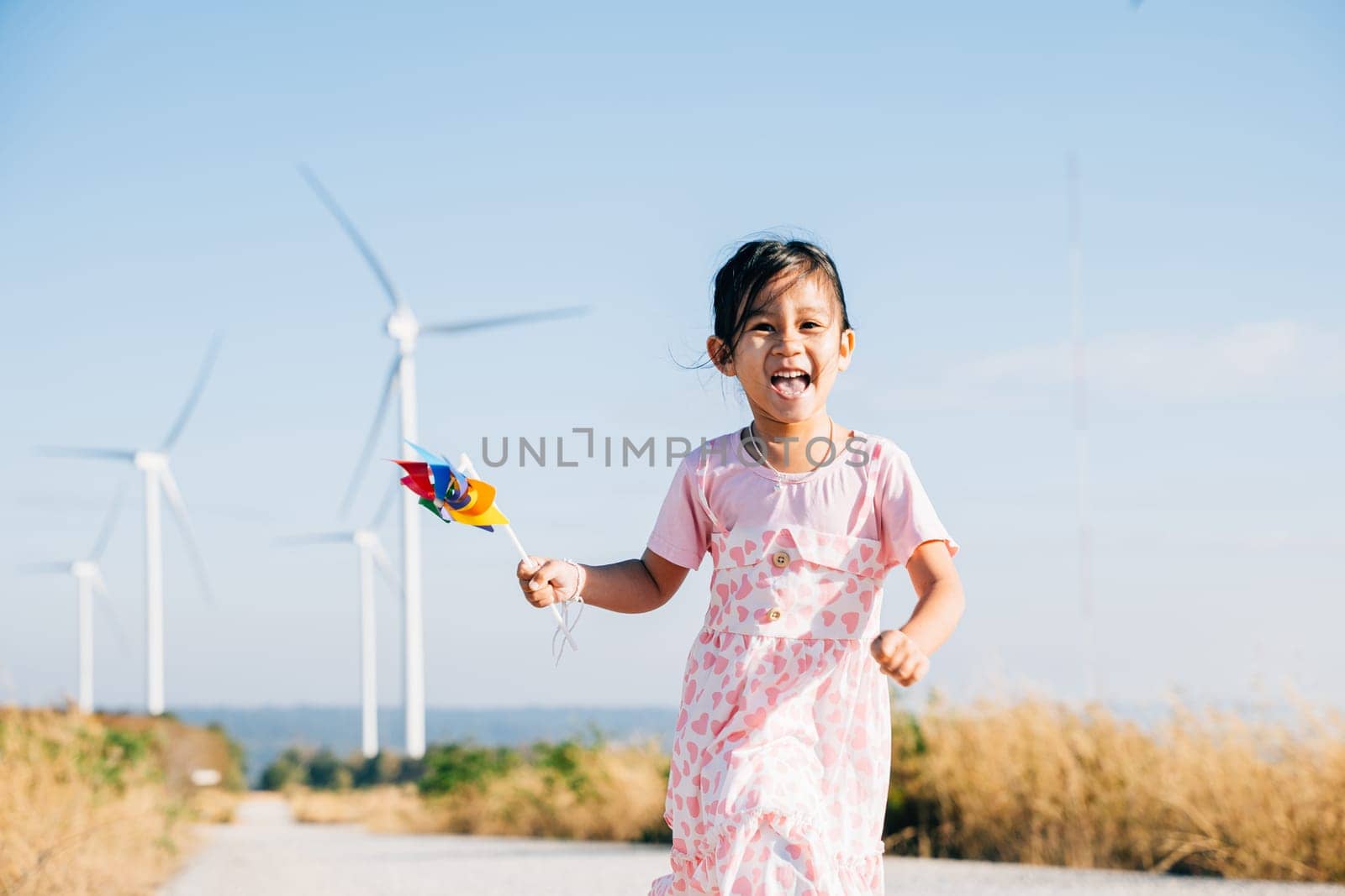 A joyful child holding pinwheels runs near windmills embodying wind energy education. Clean electricity and sustainable industry visuals in a picturesque wind turbine landscape.