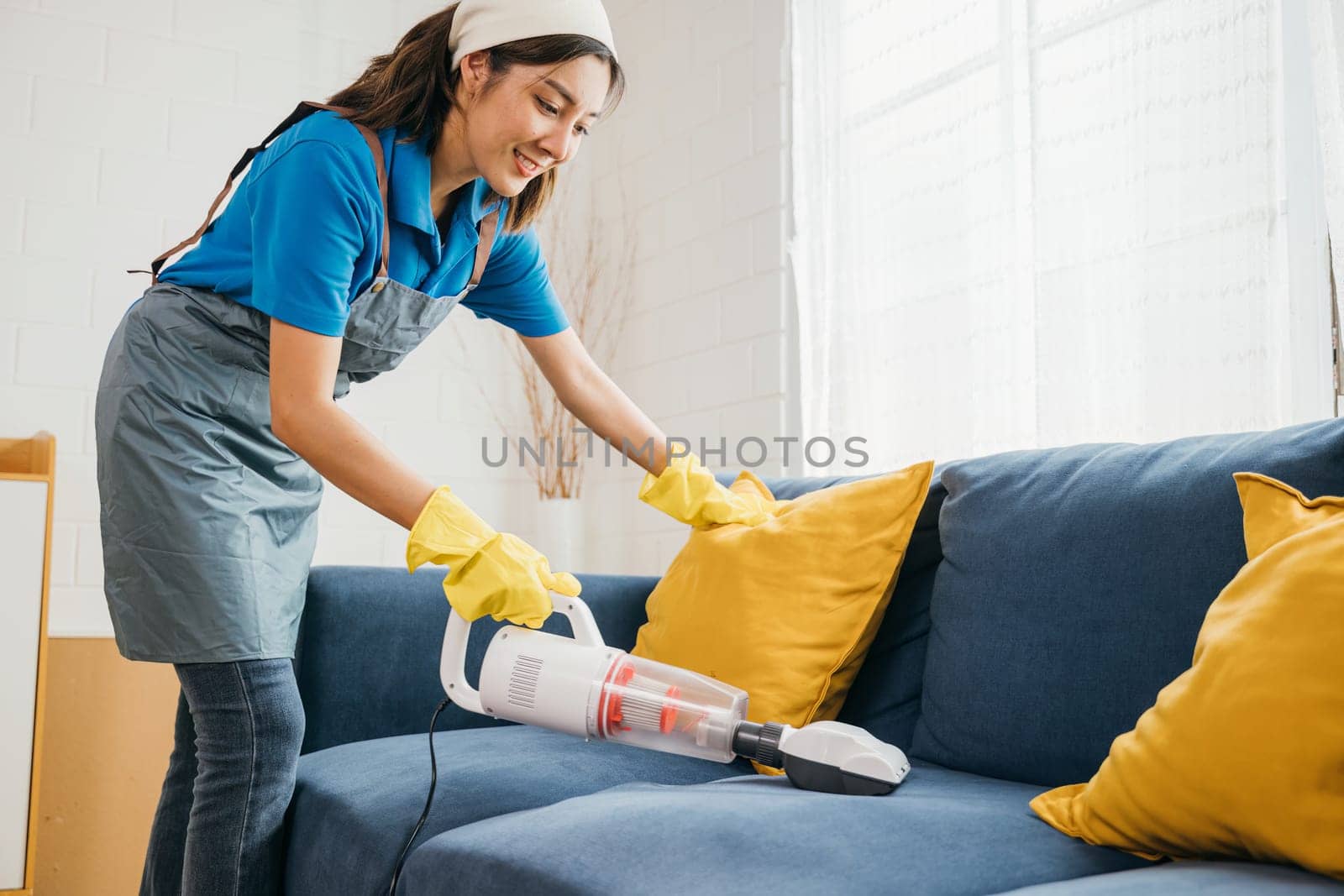 An Asian woman housekeeper employs a vacuum machine cleaner ensuring hygiene by cleaning a sofa in a living room. Her dedication to housework and furniture care reflects a modern approach. by Sorapop