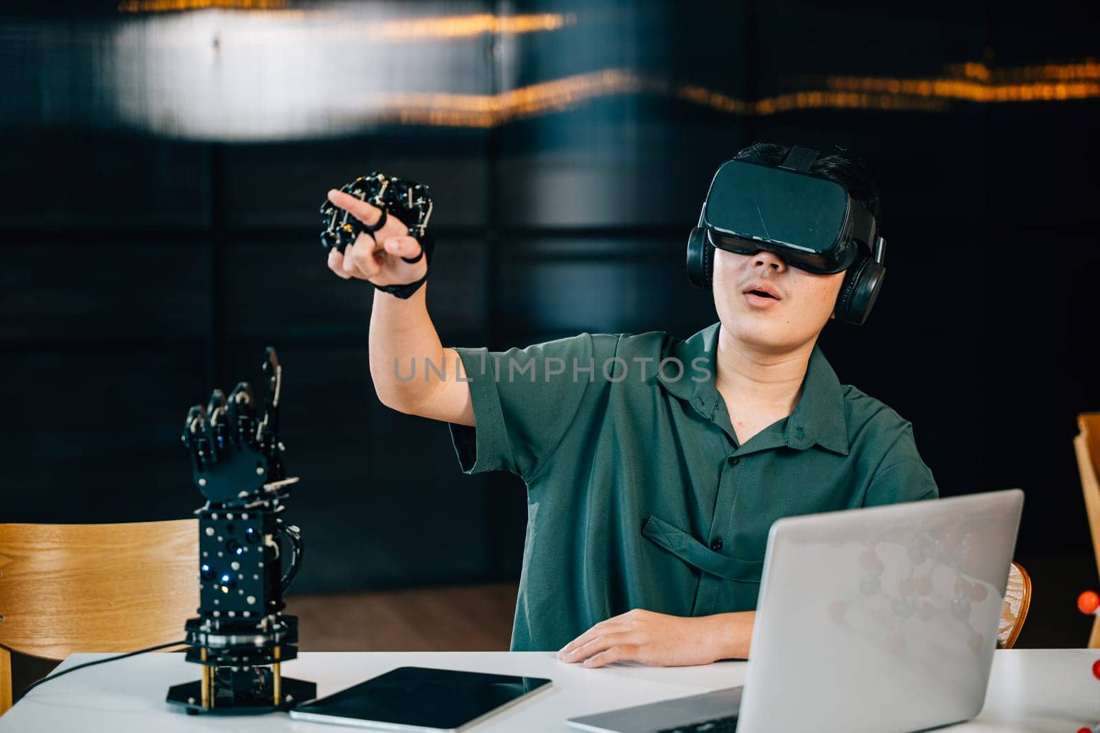 Asian teenager wears VR glasses controlling a robot arm project in a classroom. Embracing robotics programming blending technology and STEM education for futuristic learning. moves a robotic hand by Sorapop