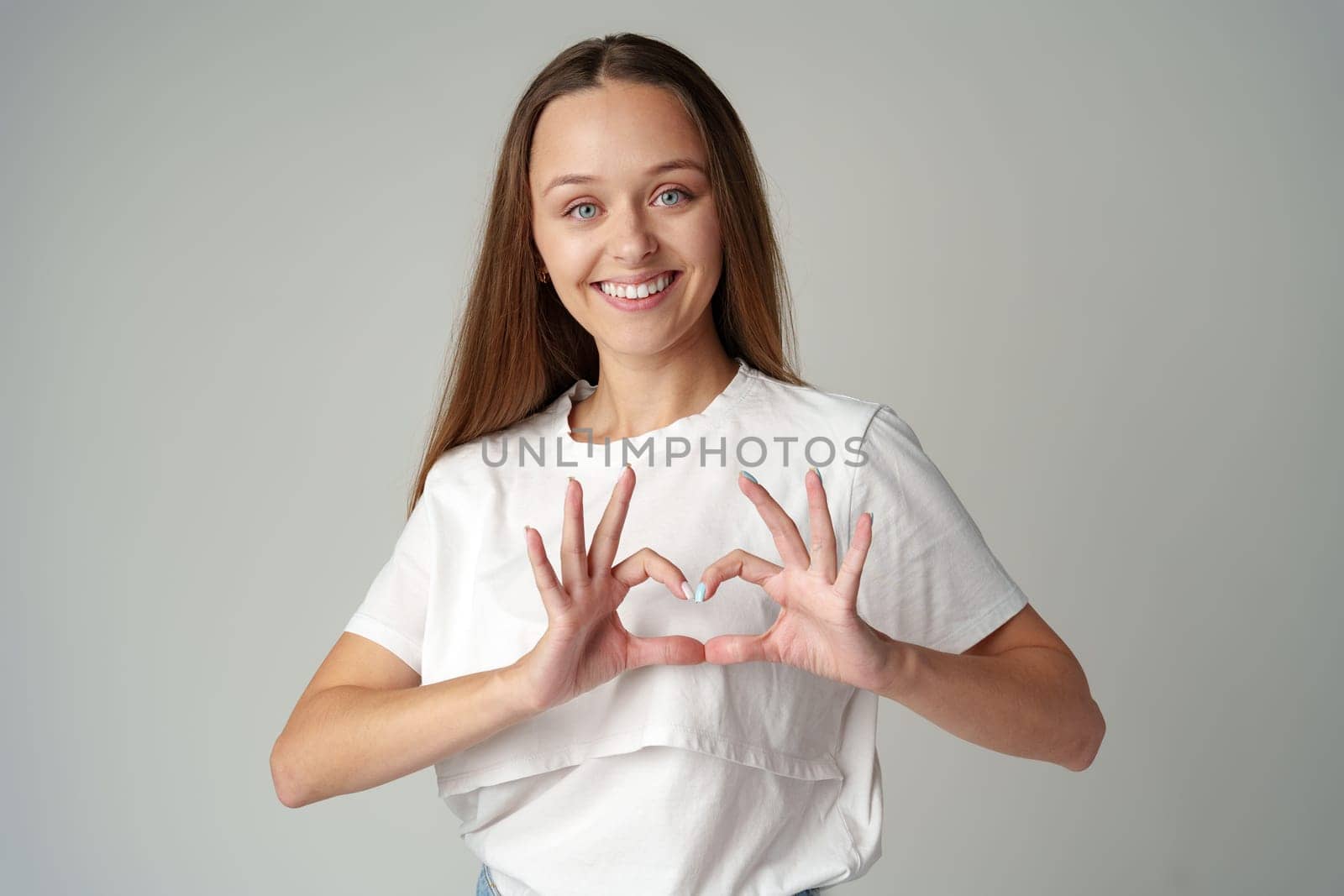 Smiling young woman showing heart sign with hands on gray background by Fabrikasimf