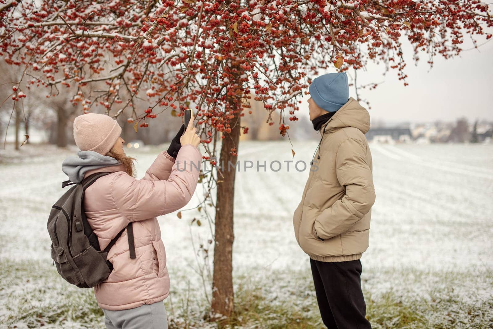 Winter Romance: Girl in Pink Winter Jacket Photographing Boy Against Snow-Covered Red Tree and Field. by Andrii_Ko