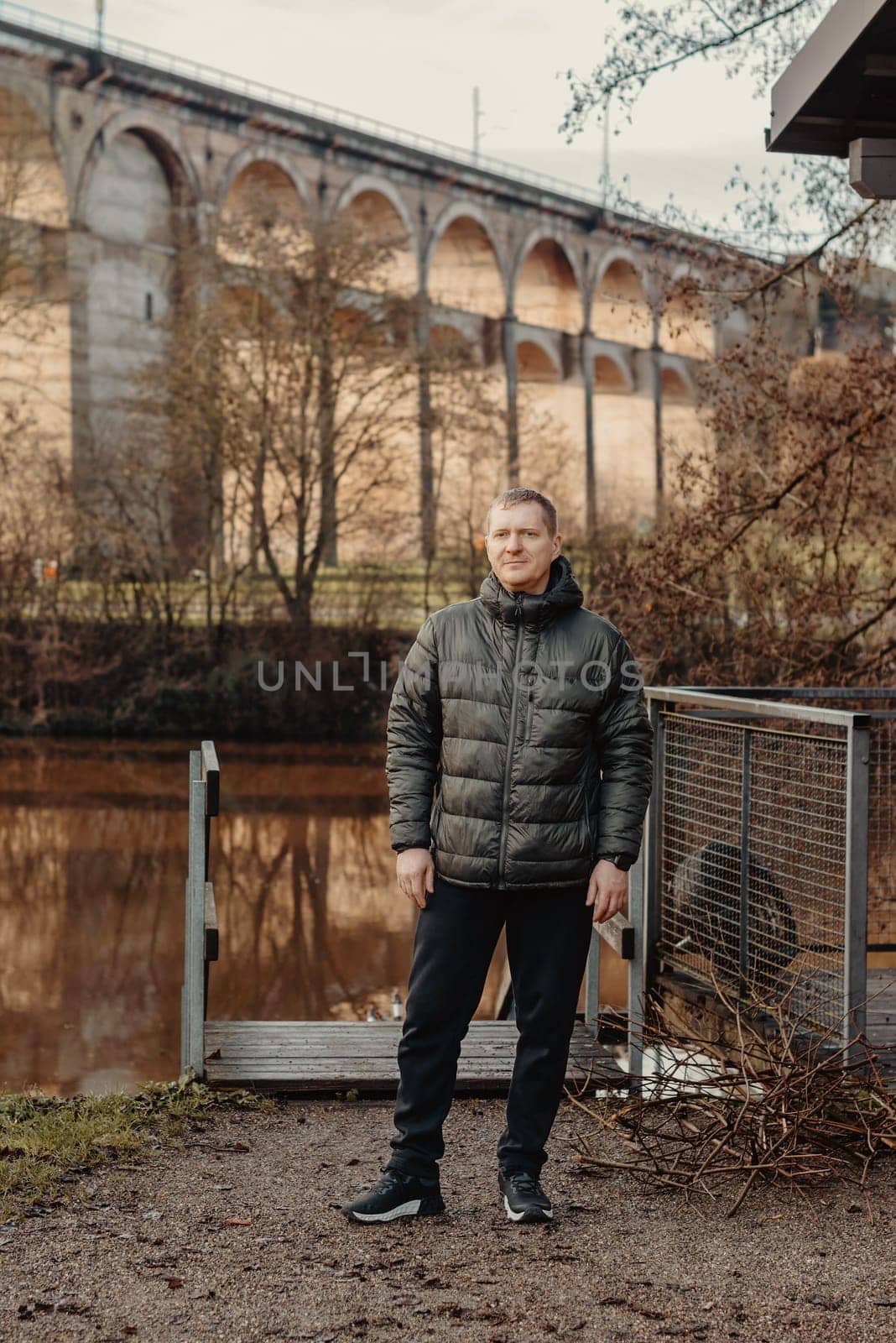 Timeless Elegance: 40-Year-Old Man in Stylish Jacket by Neckar River and Historic Bridge in Bietigheim-Bissingen, Germany. Experience the allure of seasons as a charismatic 40-year-old man stands gracefully by the enchanting Neckar River, adorned in a sophisticated jacket, against the backdrop of historic bridge pillars in the charming city of Bietigheim-Bissingen, Germany. This captivating image seamlessly blends timeless elegance with the scenic beauty of autumn or winter, creating a picturesque scene of urban exploration and mature style in the heart of German heritage