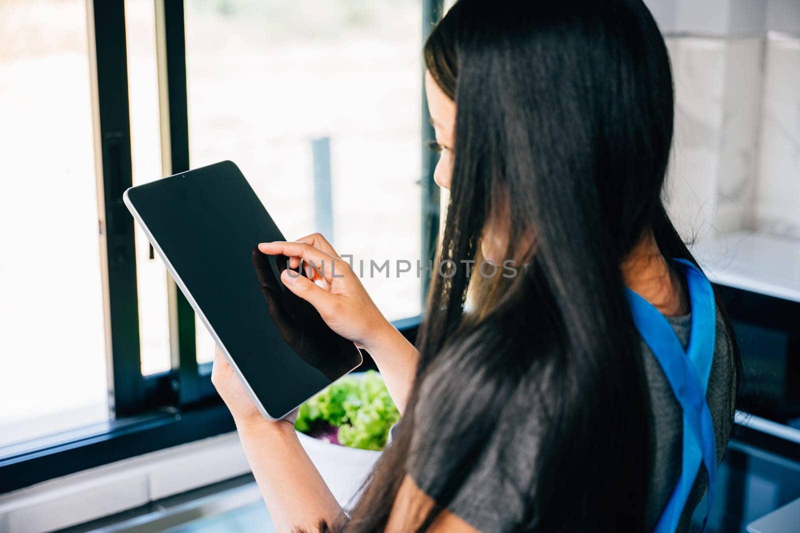 Asian woman joyfully cooks in her sunlit kitchen utilizing a tablet by Sorapop