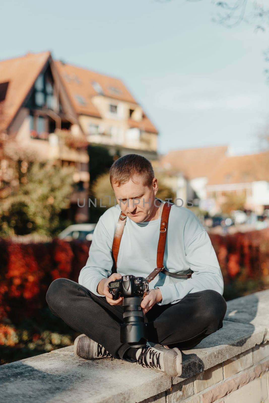 Man Sitting in Old European City And Holding Photo Camera. Contemporary Stylish Blogger And Photographer. Handsome man taking a selfie on a trip in Europe. by Andrii_Ko
