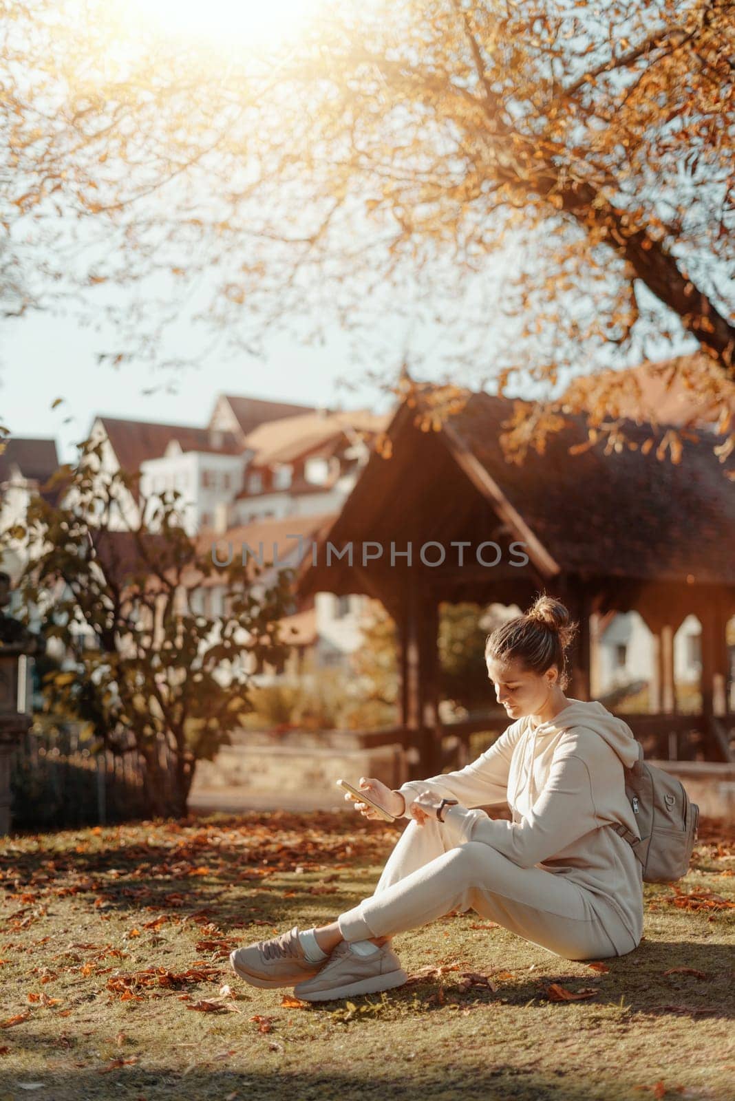 Young Fashionable Teenage Girl With Smartphone In Europian Park In Autumn Sitting At Smiling. Trendy Young Woman In Fall In Park Texting. Retouched, Vibrant Colors. Beautiful Blonde Teenage Girl Wearing Casual Modern Autumn Outfit Sitting In Park In Autumn. Retouched, Vibrant Colors, Brownish Tones. by Andrii_Ko