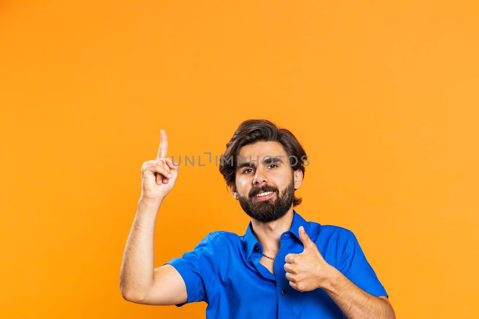 Young man showing thumbs up and pointing empty place above head, advertising area for commercial text, copy space for goods promotion advertisement. Guy isolated on orange studio background. Lifestyle