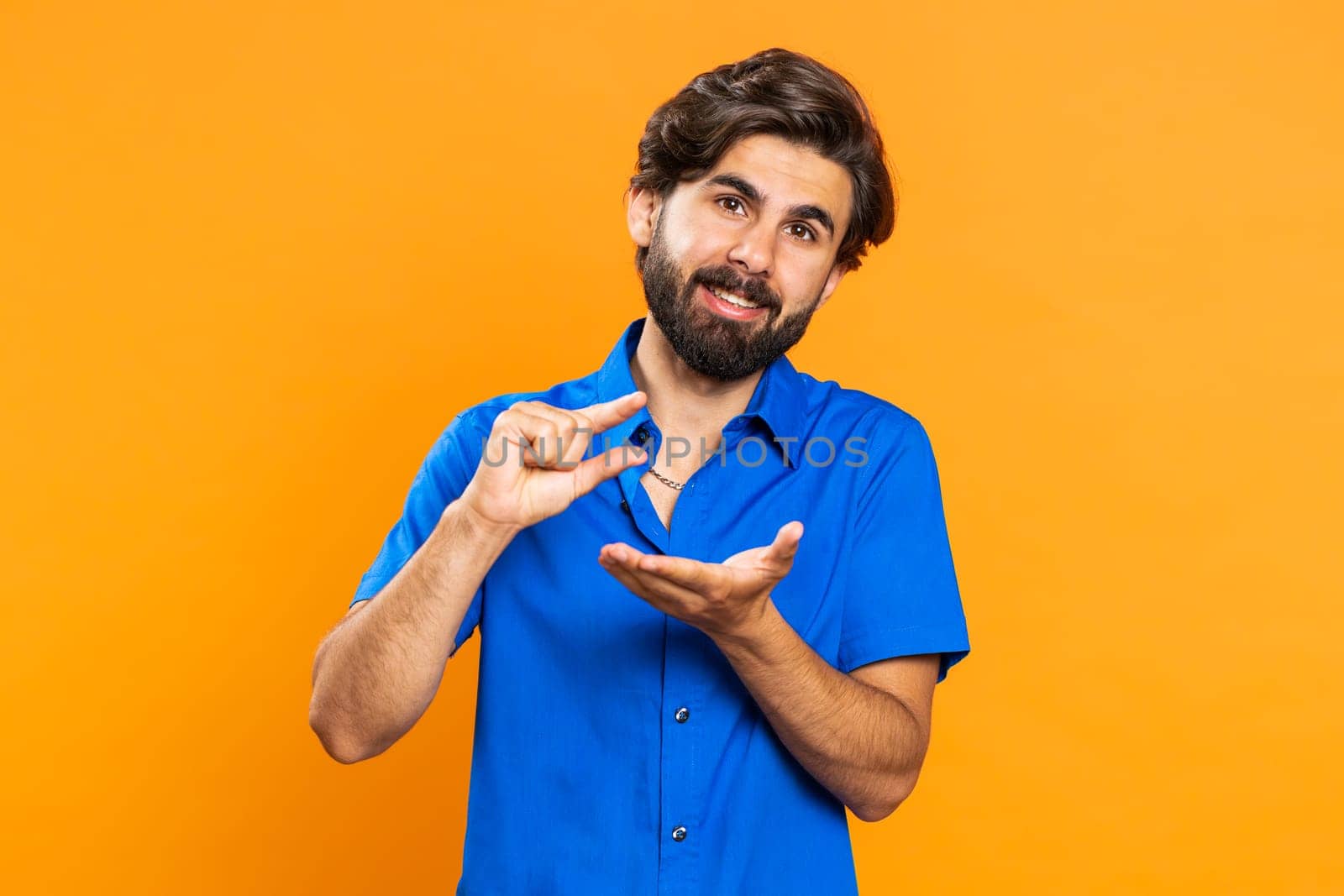 Need some more, please give me, help. Man showing a little bit gesture with sceptic smile, showing minimum sign measuring small size. Middle eastern guy isolated alone on orange studio background