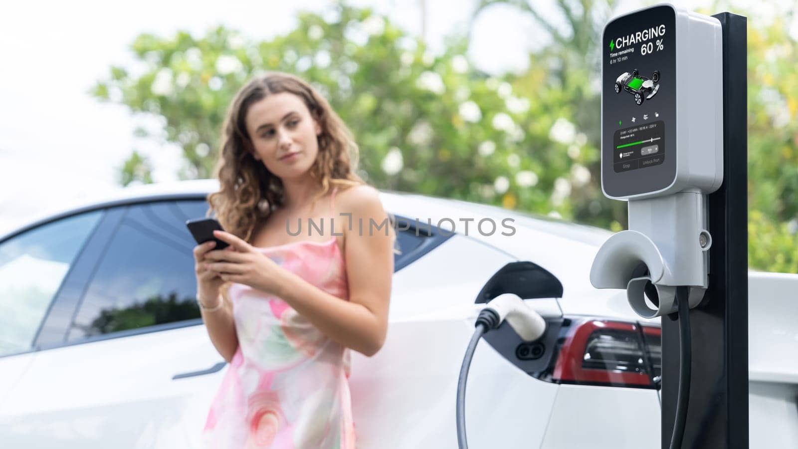 Focused charging station recharging electric vehicle on blurred background of modern woman using smartphone. EV technology utilization for tracking energy usage to optimize battery charging.Synchronos