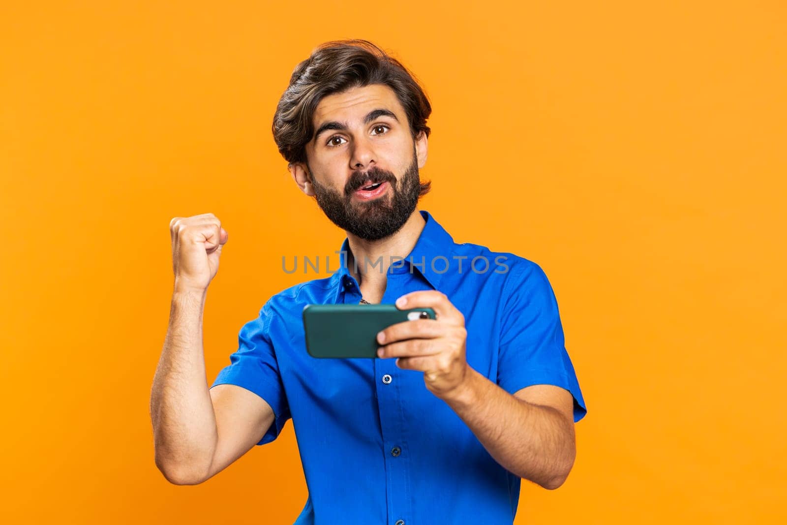 Worried funny addicted man enthusiastically playing racing video games on mobile phone. Happy middle eastern guy using smartphone gadget app with drive simulator isolated on orange studio background
