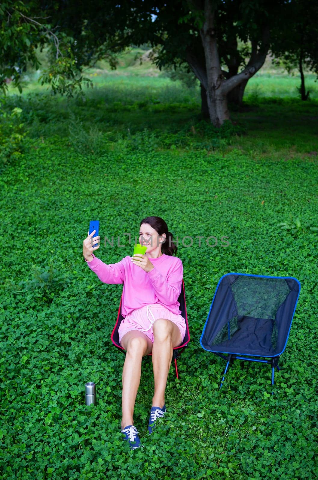 A girl in pink takes a selfie outdoors while sitting on a blue chair among lush greenery