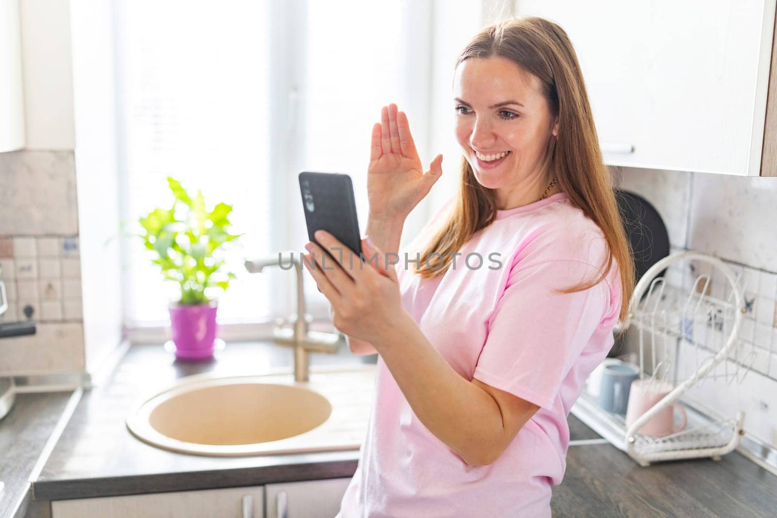 Happy woman in pink t-shirt waving while having a video call on her smartphone in a bright, sunny kitchen.