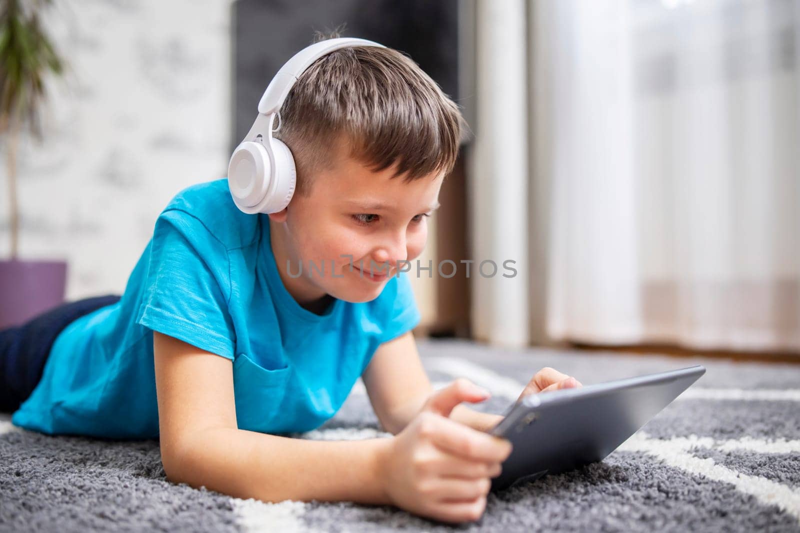 Boy lying on carpet wearing headphones and using tablet. Casual indoor leisure activity. Child technology use concept