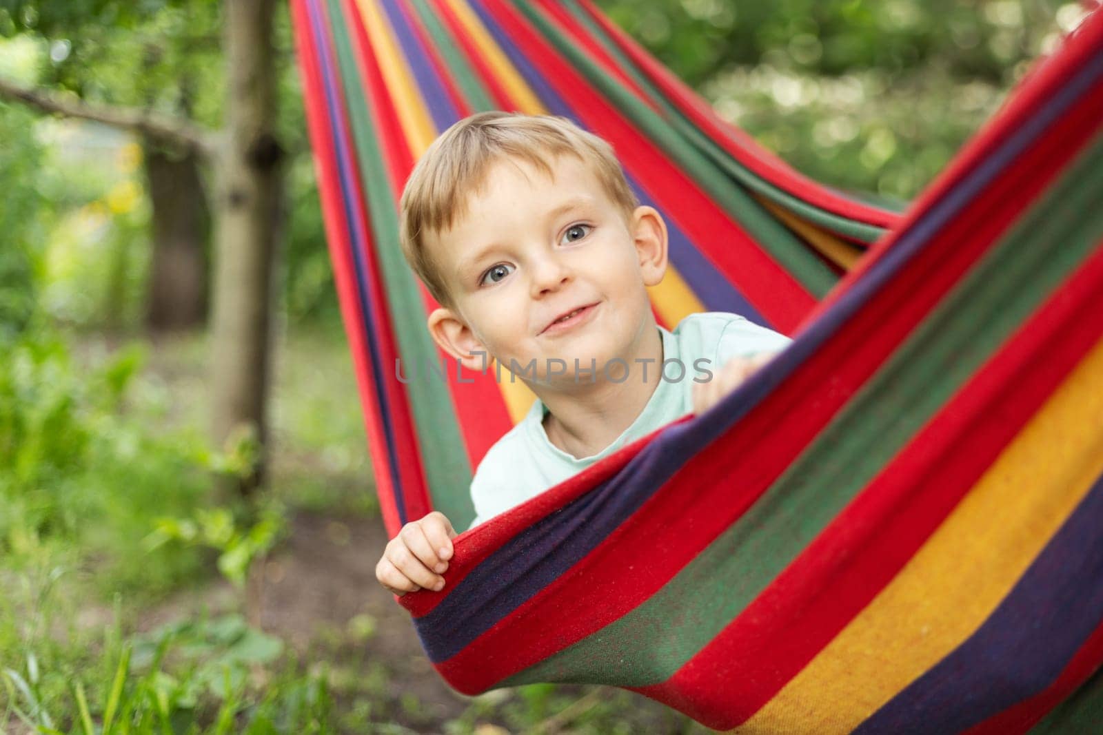 Smiling child peeking from colorful hammock. Outdoor relaxation and joy concept. Summer leisure scene. Design for greeting card, postcard