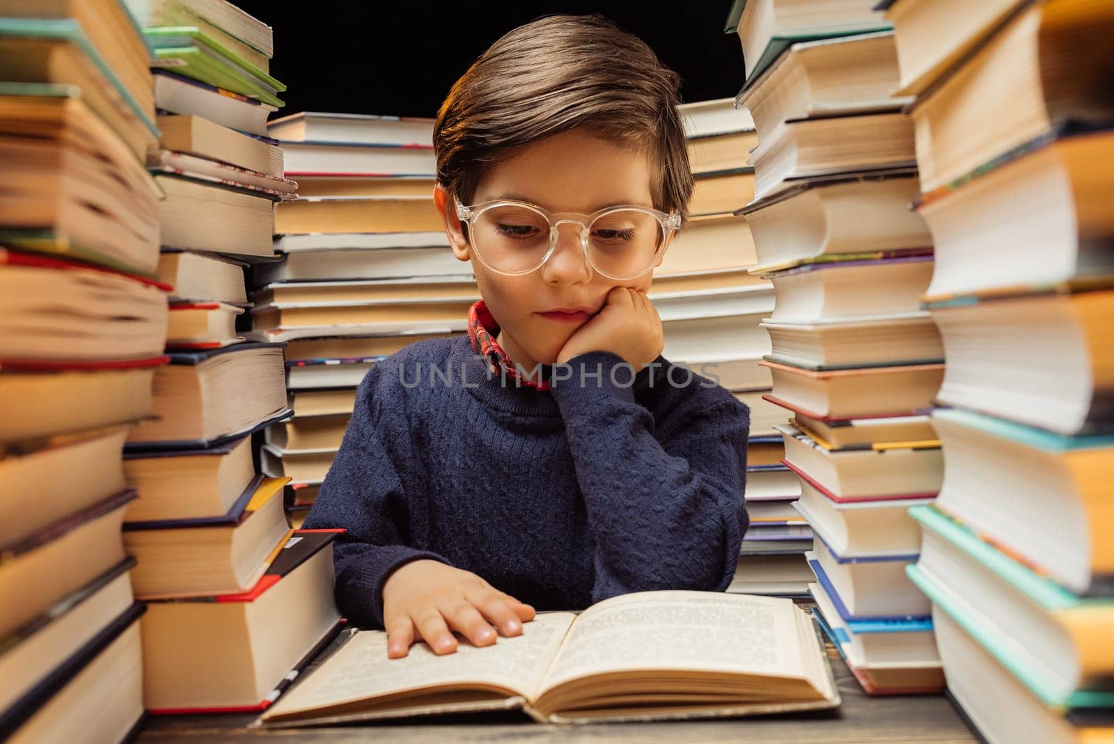 Cute little pupil boy in glasses reading interesting book in library between stacks of books literature. Education concept, prep or elementary school. High quality