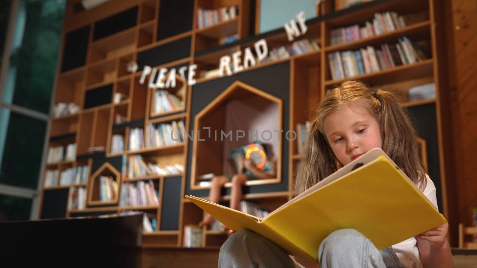 Young smart caucasian girl picking reading a book while sitting at library. Clever child learning, studying, open a books at library. Attractive kid turning page with blurring background. Erudition.