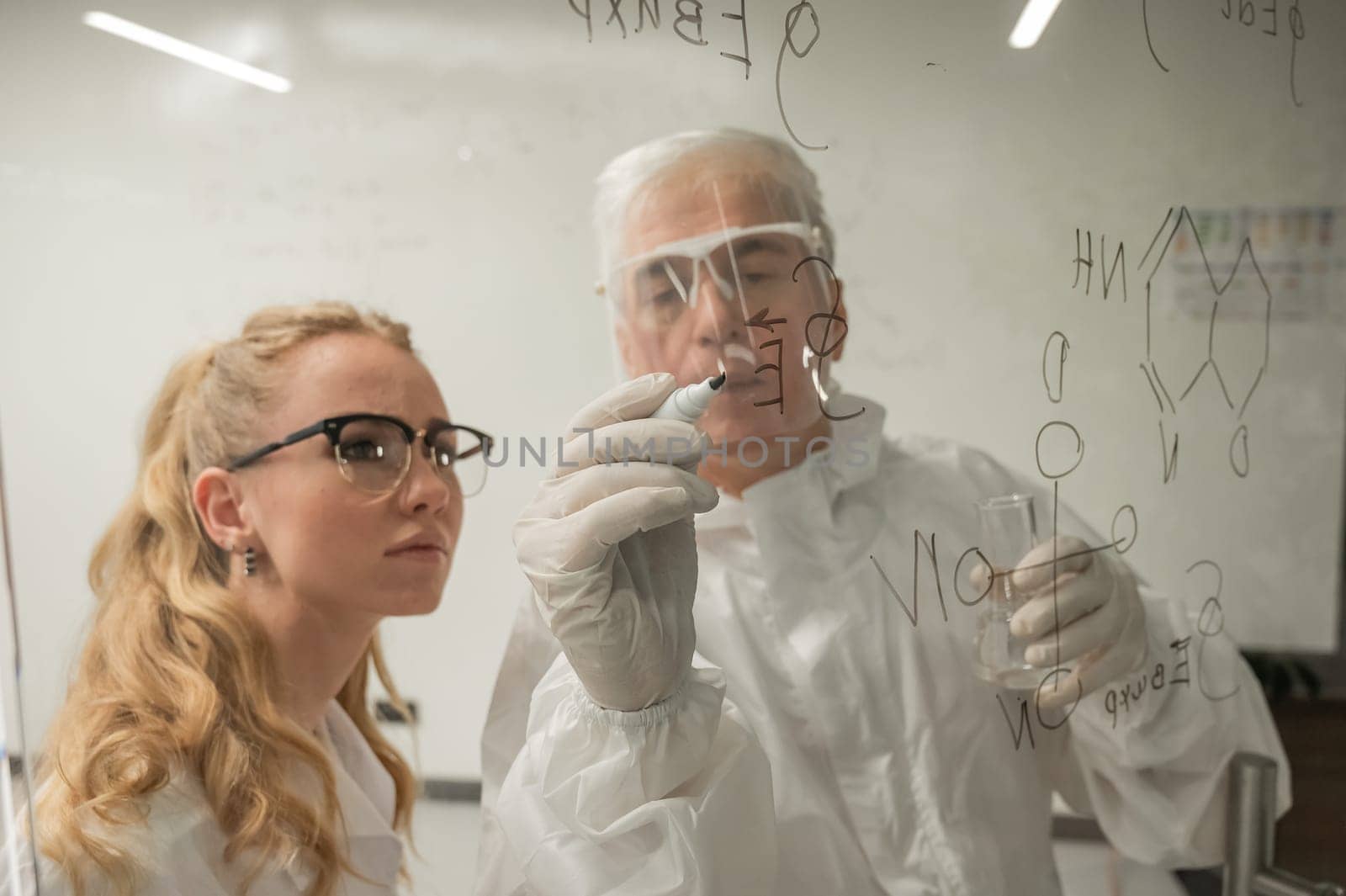 Two chemist colleagues write formulas on glass. Caucasian elderly man and young woman brainstorming. by mrwed54
