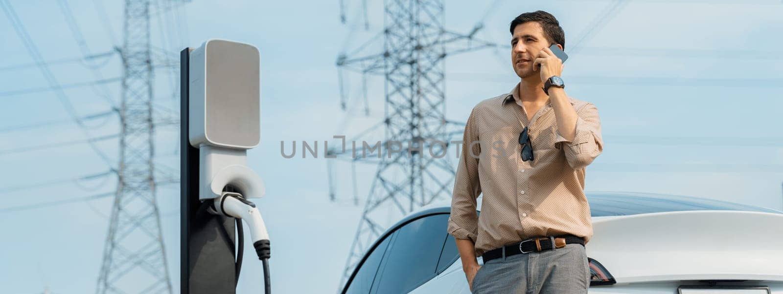 Man talking on the phone while recharge EV car battery at charging station connected to power grid tower electrical as electrical industry for eco friendly car utilization. Panorama Expedient