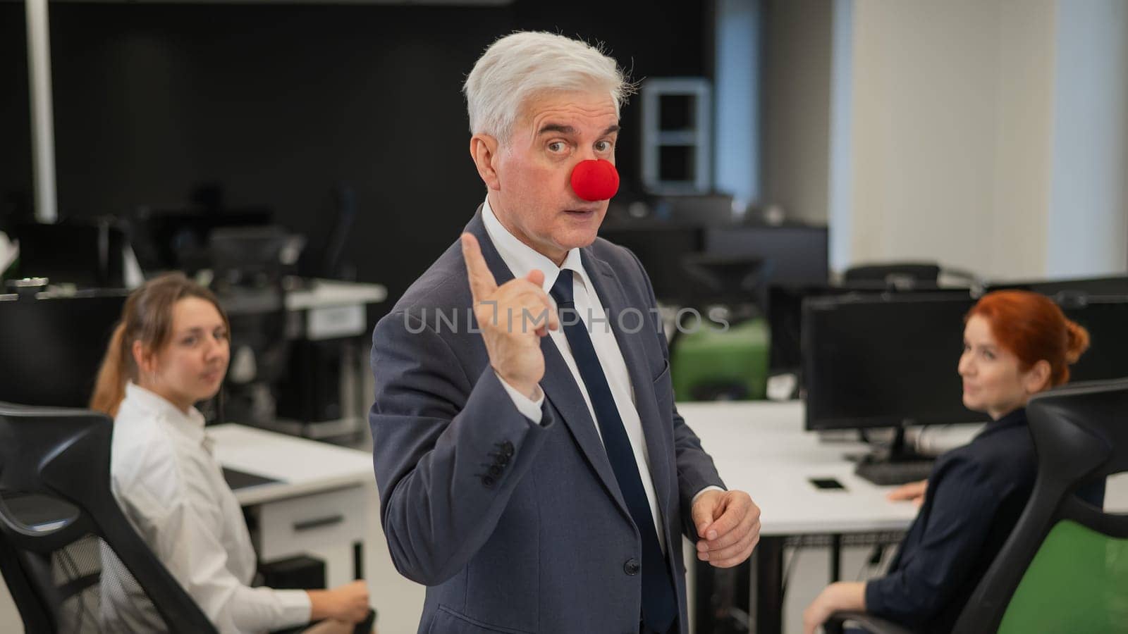 An elderly man with a clown nose stands next to two Caucasian women in the office