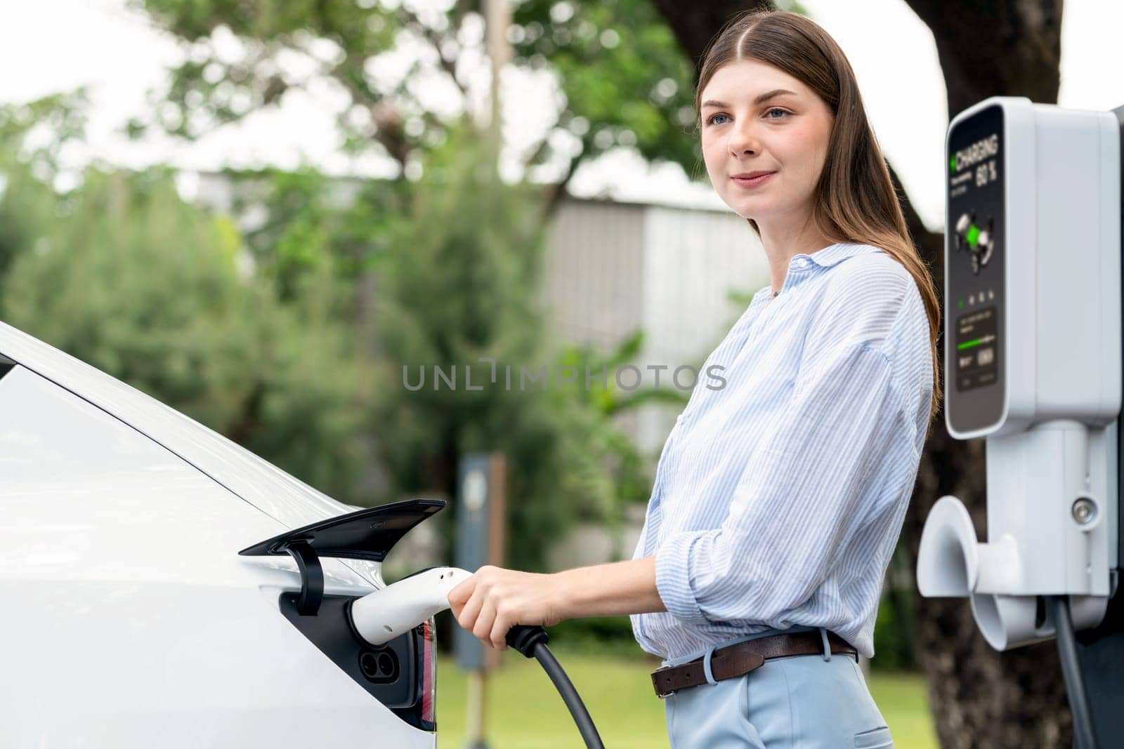 Young woman recharge EV electric vehicle's battery from EV charging station in outdoor green city park scenic. Eco friendly urban transport and commute with eco friendly EV car travel. Exalt