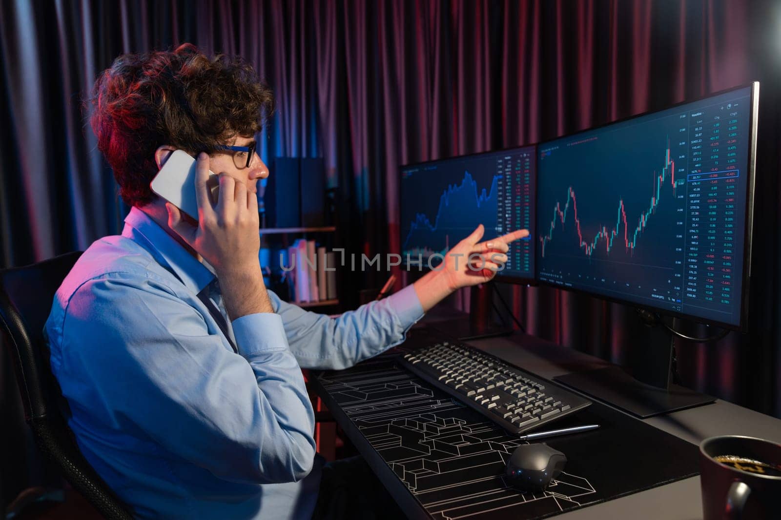 Working young business trader discussing with broker on smartphone with market stock graph in real time monitor screens at neon light modern office. Concept of analyzing dynamic exchange rate. Gusher.