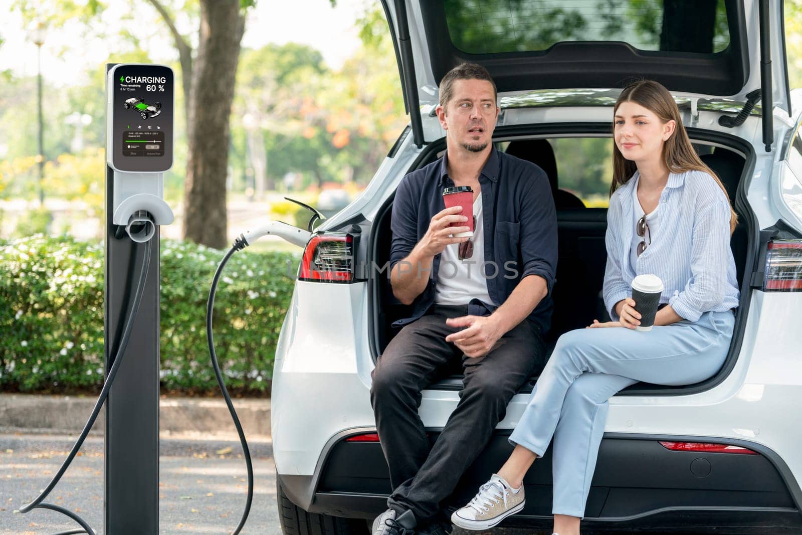 Lovely young couple drinking coffee while recharging battery for electric car during road trip travel EV car in natural forest or national park. Eco friendly travel during vacation and holiday. Exalt