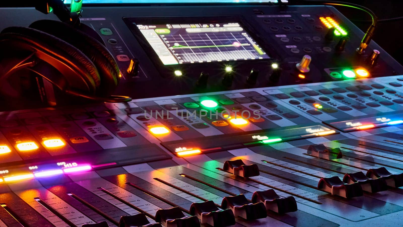 Low angle close up on a dark studio mixing sound board, with rows of small dials illuminated by a rainbow spot light, in the control booth at a concert