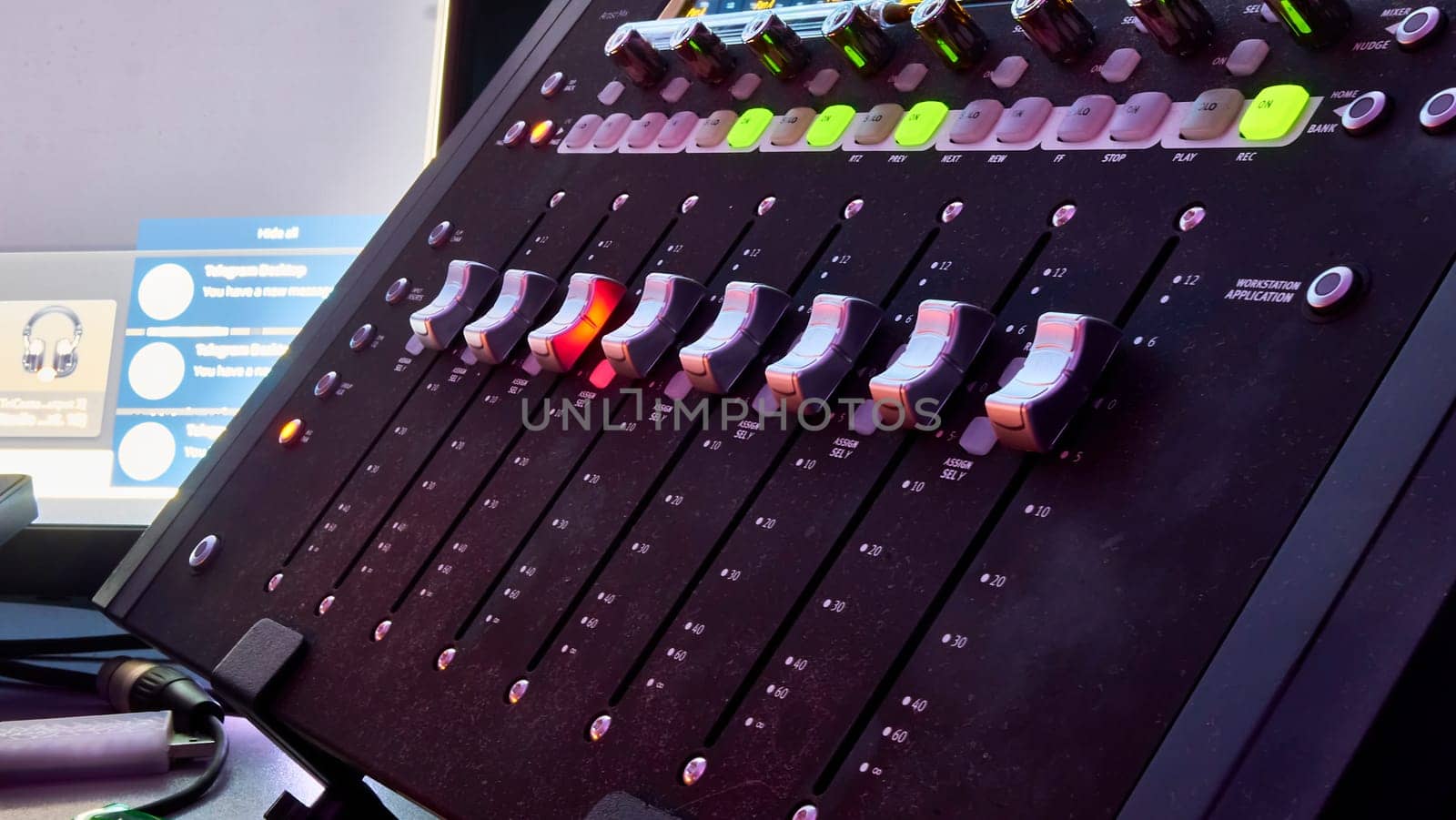 Low angle close up on a dark studio mixing sound board, with rows of small dials illuminated by a rainbow spot light, in the control booth at a concert