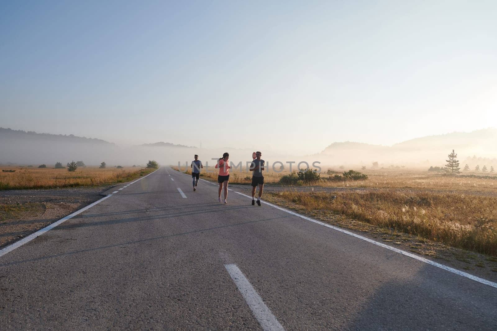 A group of friends, athletes, and joggers embrace the early morning hours as they run through the misty dawn, energized by the rising sun and surrounded by the tranquil beauty of nature.