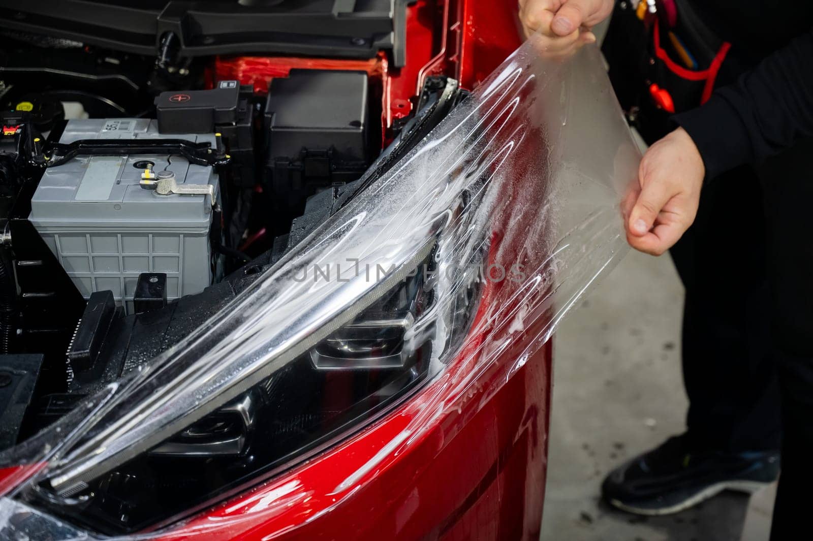 The master applies vinyl film to the headlight of a red car. Closeup view on worker detailer hand smoothing with a scraper protective film. by mrwed54