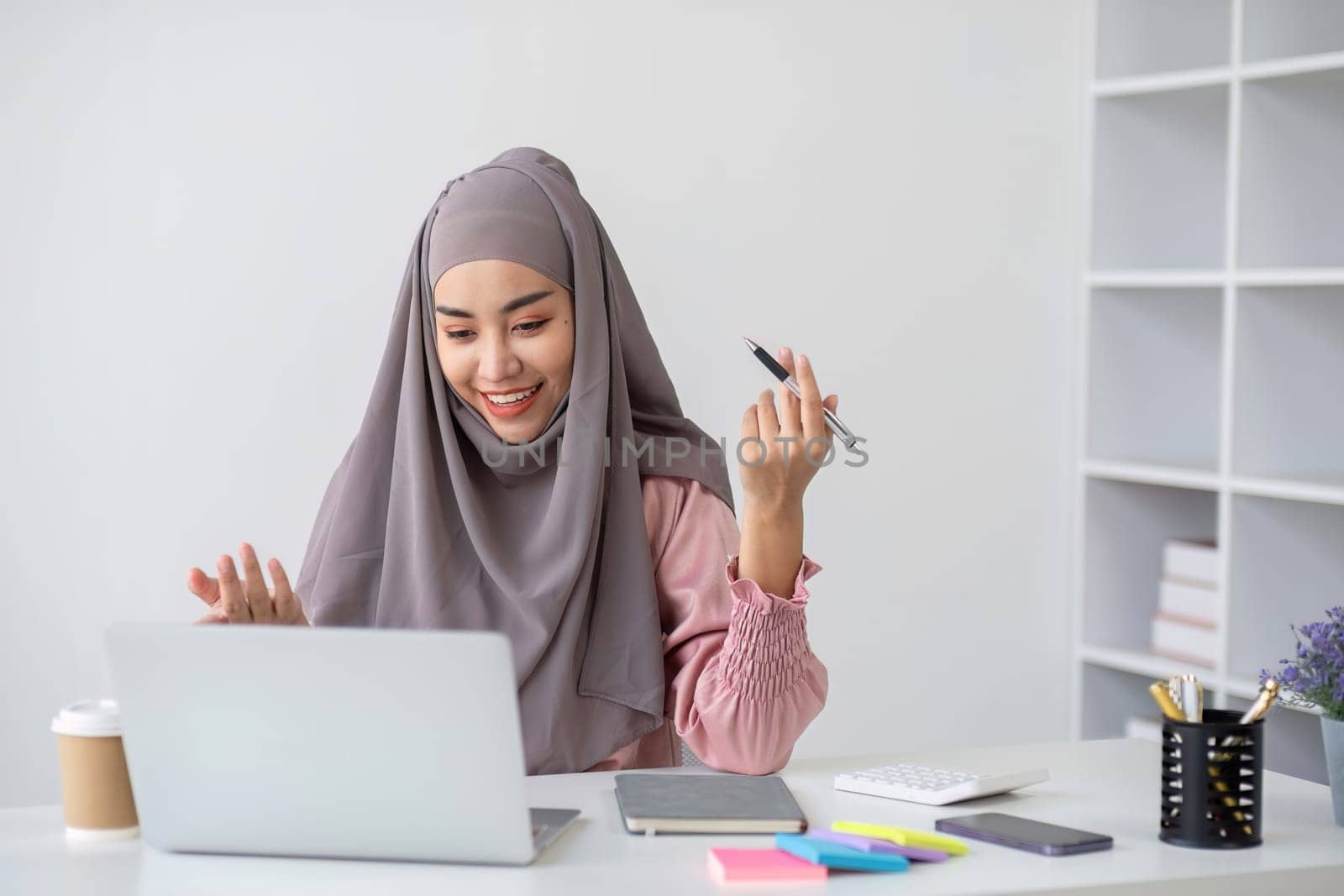 Smiling Muslim business woman Wearing a hijab while chatting online on a work desk in the office. by wichayada