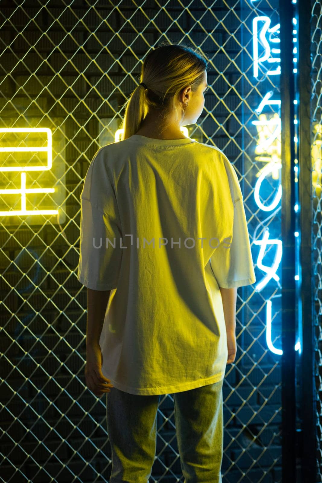 A beautiful blonde girl in a white oversized T-shirt and blue jeans posing against the background of neon hieroglyphs. High quality photo