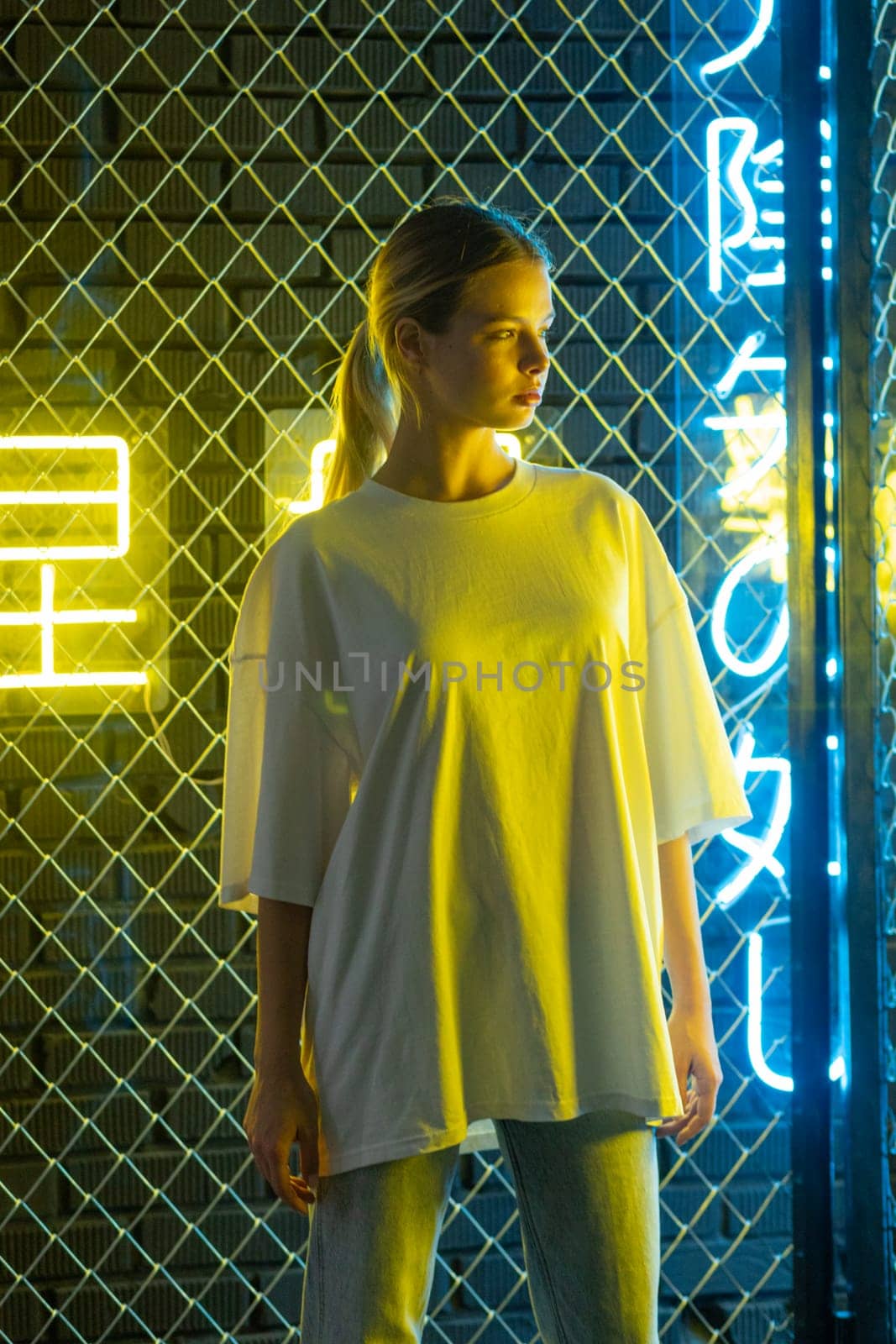 A beautiful blonde girl in a white oversized T-shirt and blue jeans posing against the background of neon hieroglyphs. High quality photo