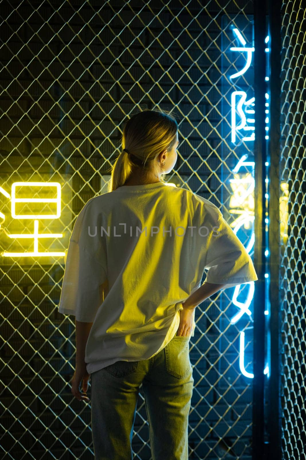 A beautiful blonde girl in a white oversized T-shirt and blue jeans posing against the background of neon hieroglyphs. High quality photo