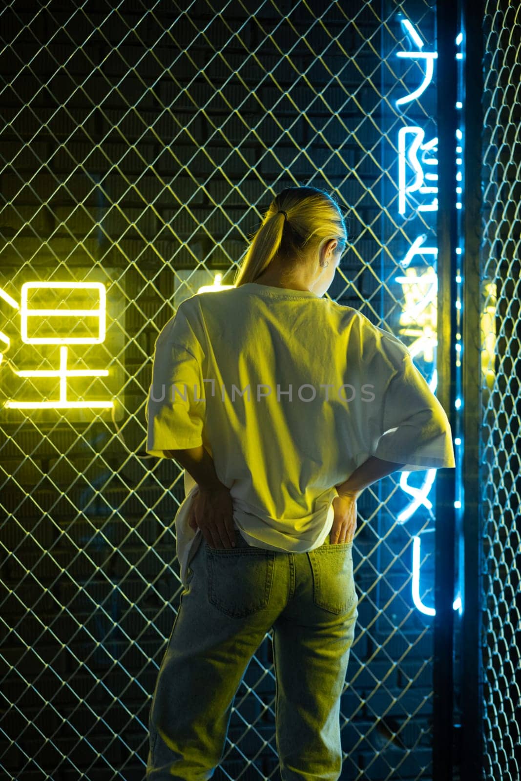 A beautiful blonde girl in a white oversized T-shirt and blue jeans posing against the background of neon hieroglyphs. High quality photo