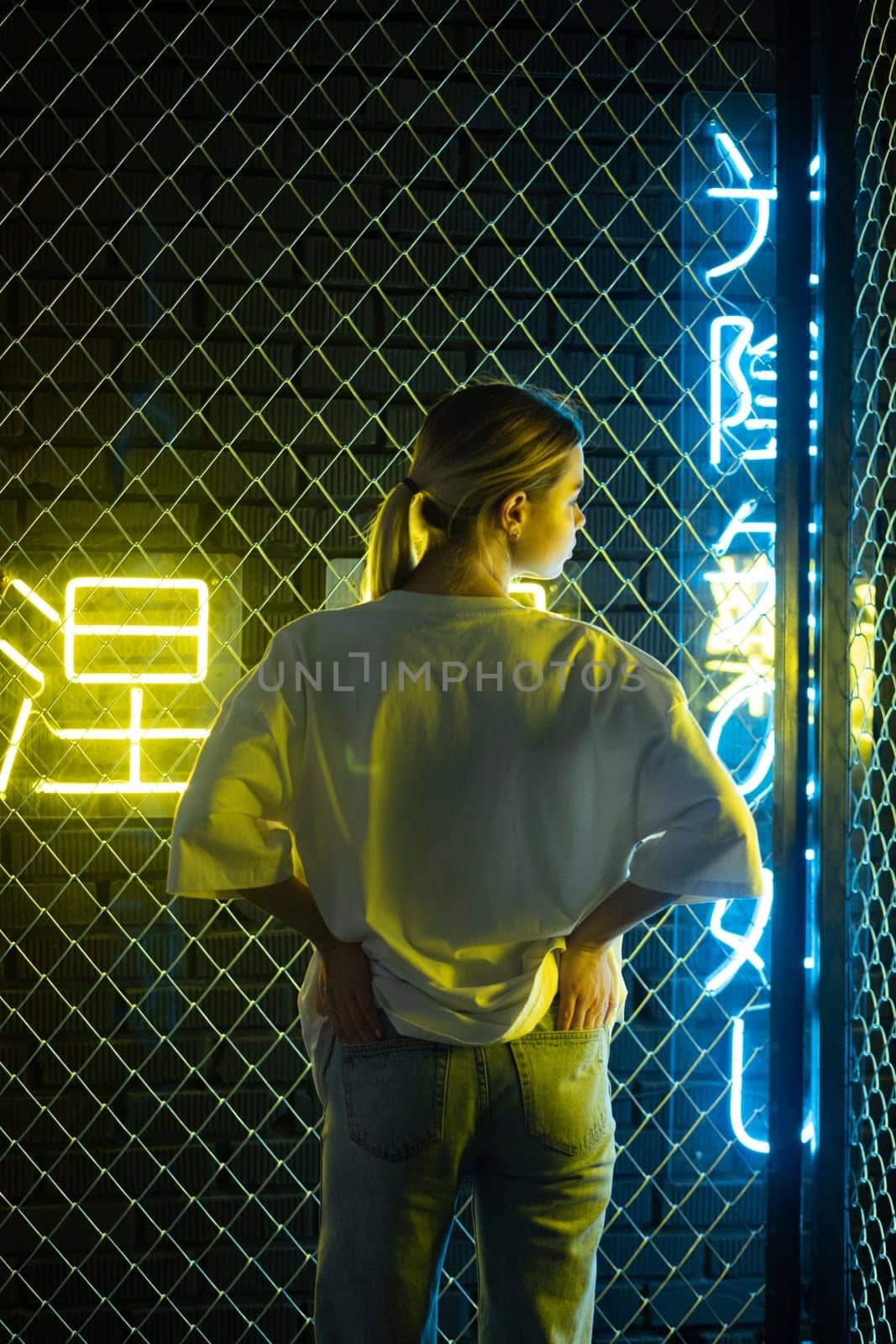 A beautiful blonde girl in a white oversized T-shirt and blue jeans posing against the background of neon hieroglyphs by Freeman_Studio