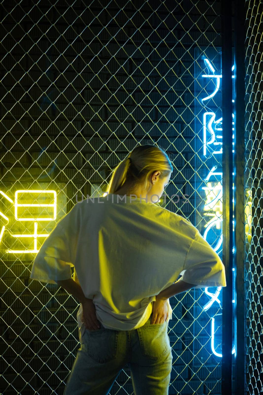 A beautiful blonde girl in a white oversized T-shirt and blue jeans posing against the background of neon hieroglyphs by Freeman_Studio
