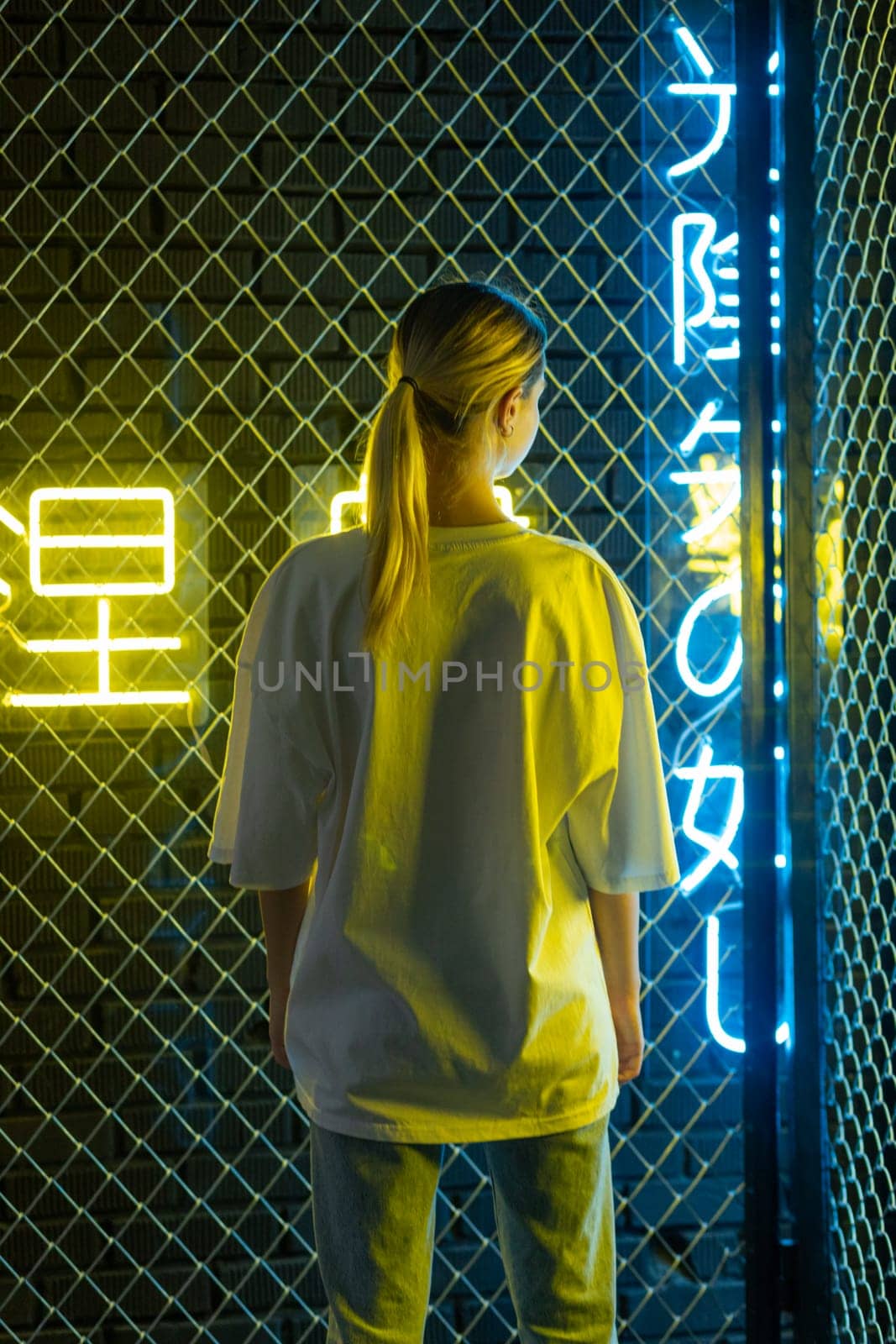 A beautiful blonde girl in a white oversized T-shirt and blue jeans posing against the background of neon hieroglyphs. High quality photo