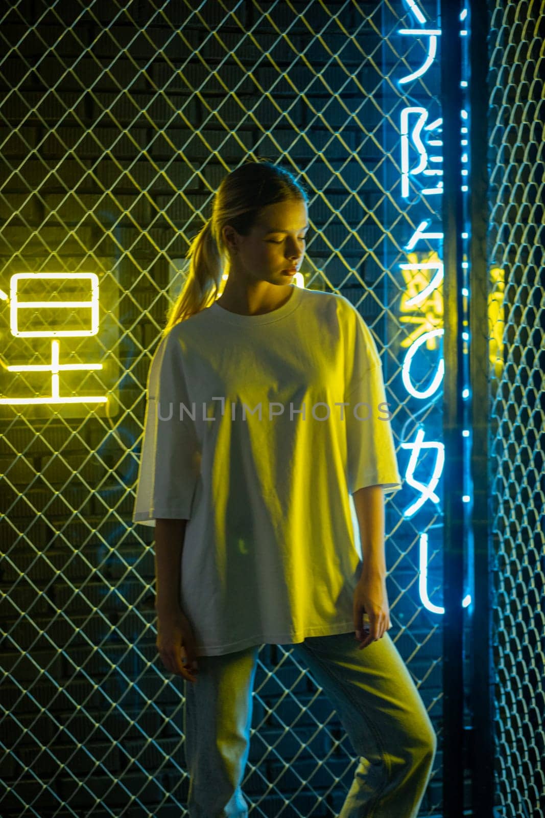 A beautiful blonde girl in a white oversized T-shirt and blue jeans posing against the background of neon hieroglyphs. High quality photo