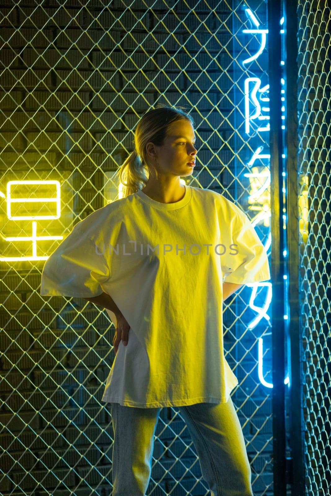 A beautiful blonde girl in a white oversized T-shirt and blue jeans posing against the background of neon hieroglyphs by Freeman_Studio