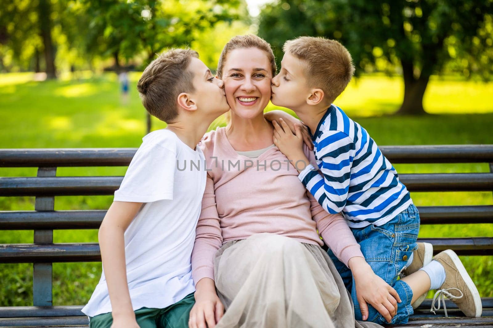 Happy mother is sitting with her sons on bench in park. They are having fun together. Boys are kissing their mother.