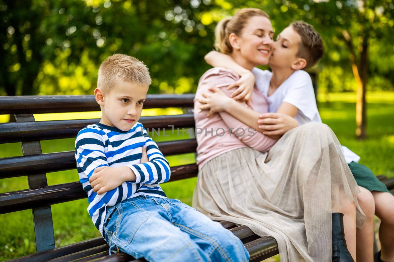 Happy mother is sitting with her sons on bench in park. One boy is offended and pouting.