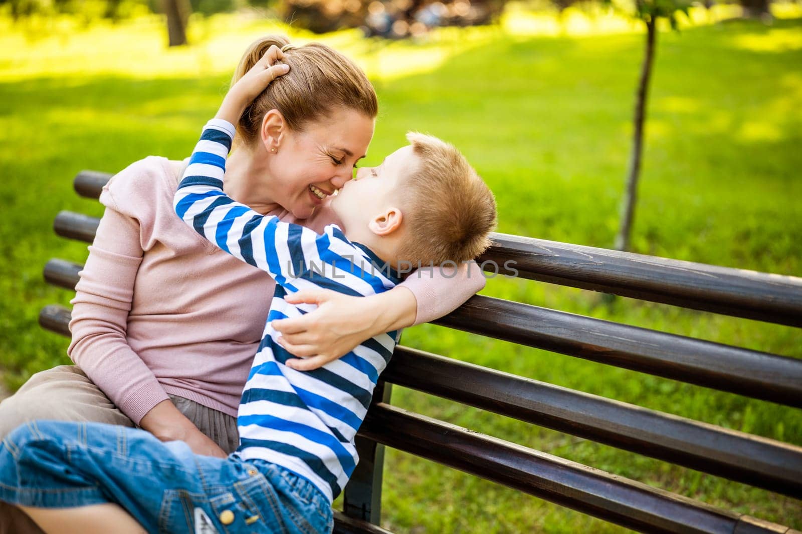 Happy mother is sitting with her son on bench in park. They are having fun together.