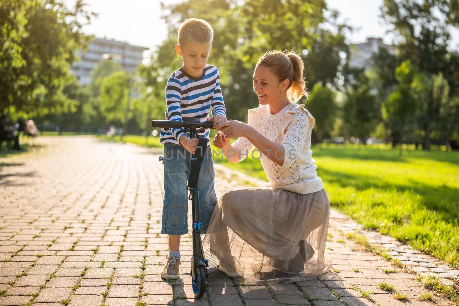 Mother having fun with her son in park on sunny day. Boy is ridding scooter.