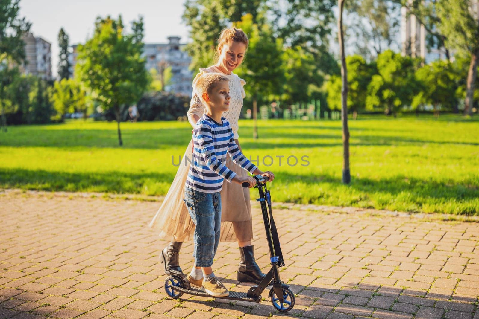 Mother having fun with her son in park on sunny day. Boy is ridding skateboard and his mother is pushing him.