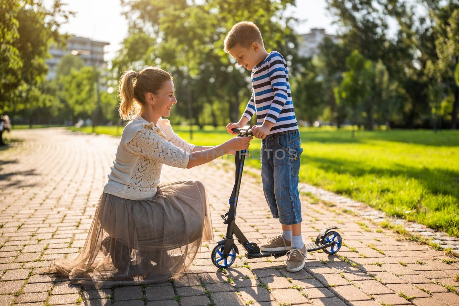 Mother having fun with her son in park on sunny day. Boy is ridding scooter.