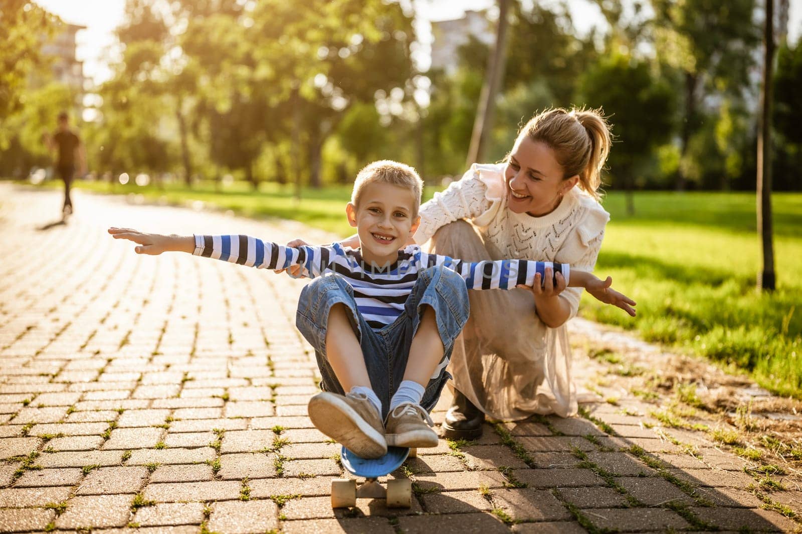 Mother having fun with her son in park on sunny day. Boy is ridding skateboard and his mother is pushing him.