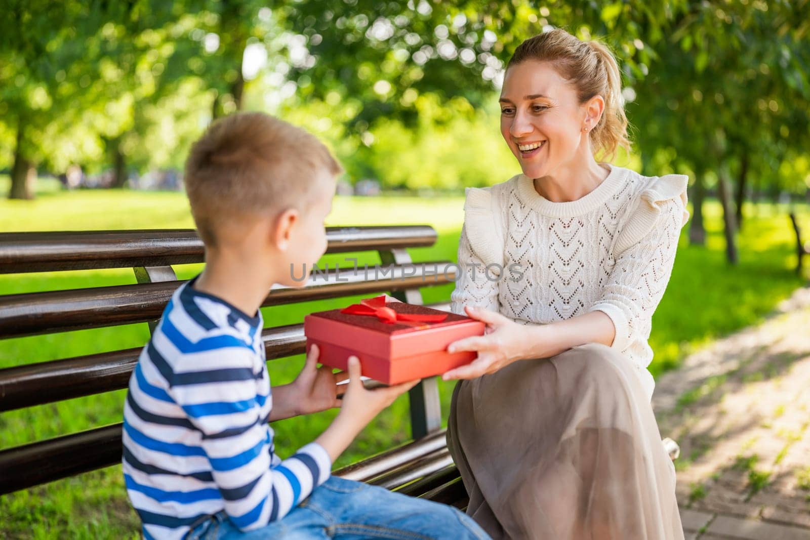Happy mother is sitting with her son on bench in park. Boy is giving a present to his mother.