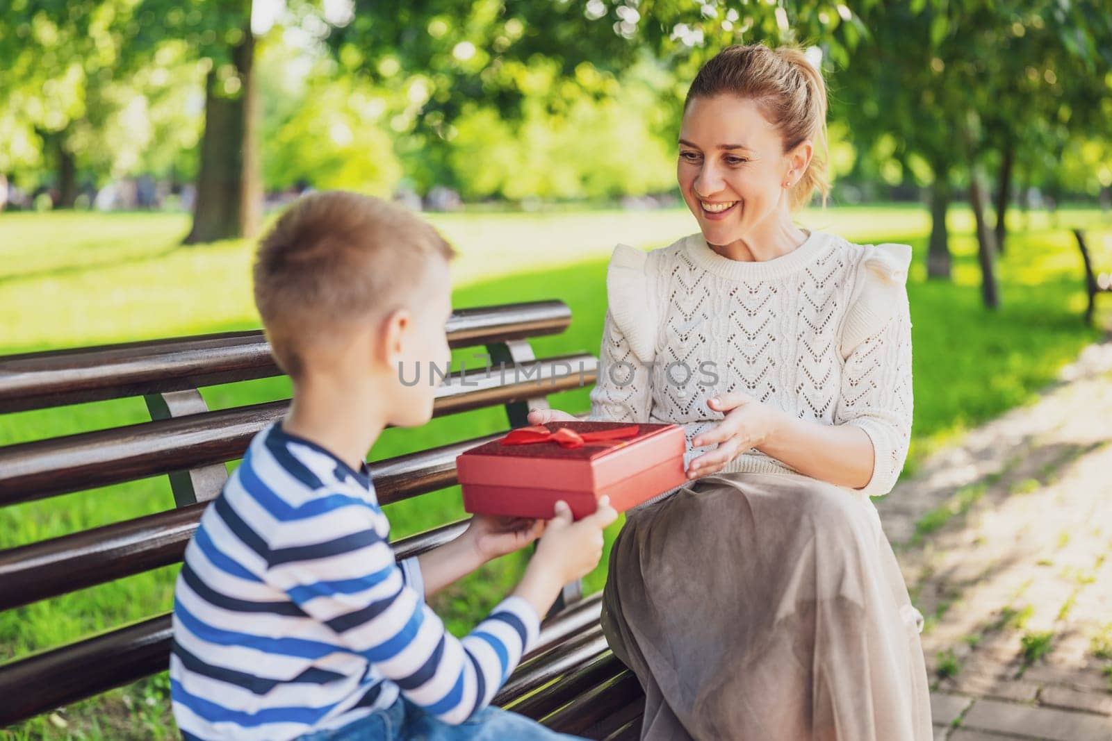 Happy mother is sitting with her son on bench in park. Boy is giving a present to his mother.