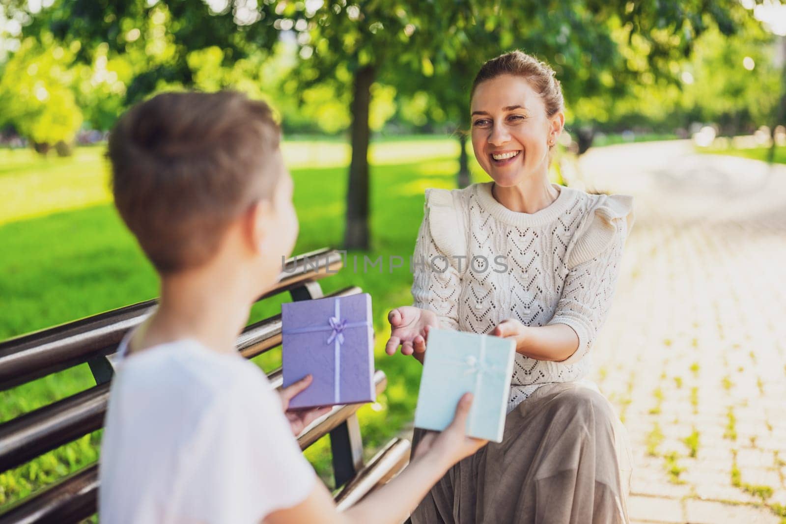 Happy mother is sitting with her son on bench in park. Boy is giving a present to his mother.