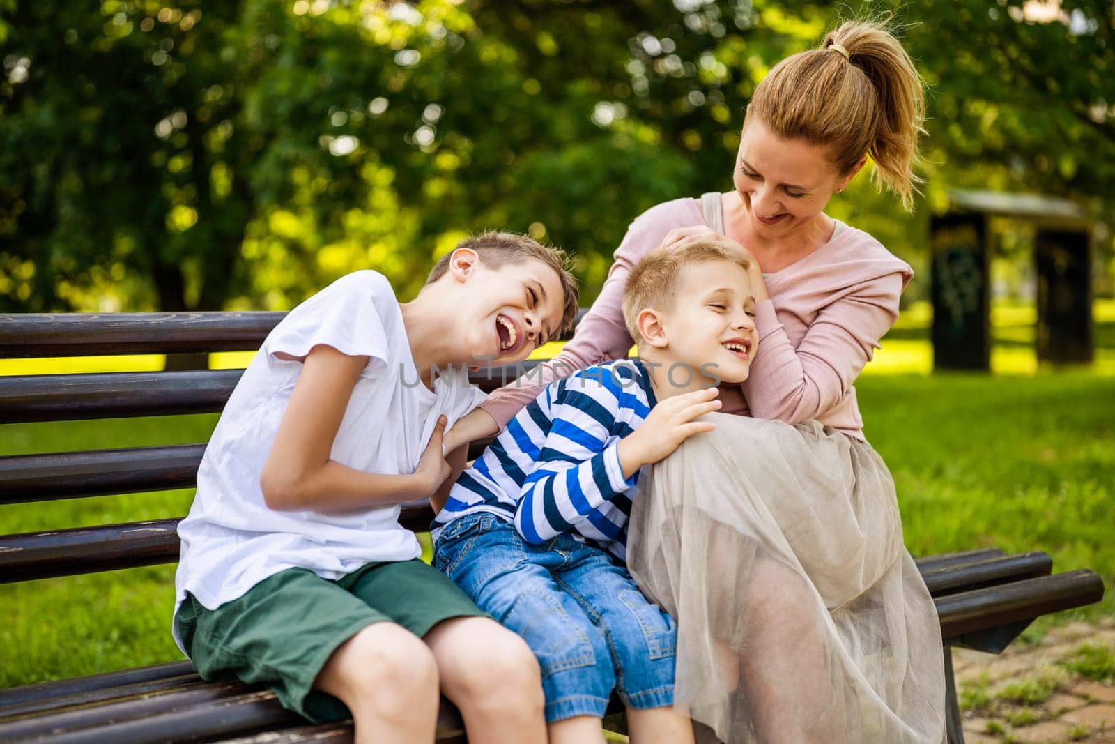 Happy mother is sitting with her sons on bench in park. They are talking and enjoying their time together.